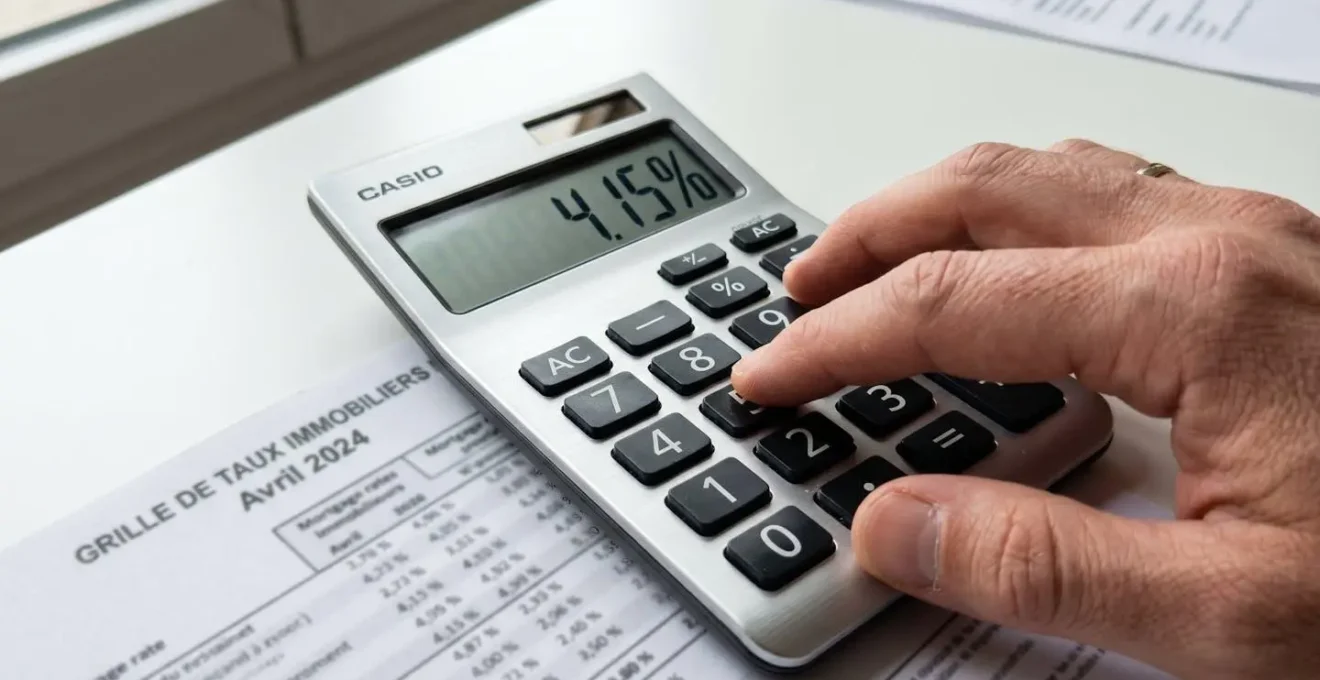 Gros plan sur une main utilisant une calculatrice moderne posée à côté d'une feuille de taux de crédit immobilier, avec un arrière-plan flouté
