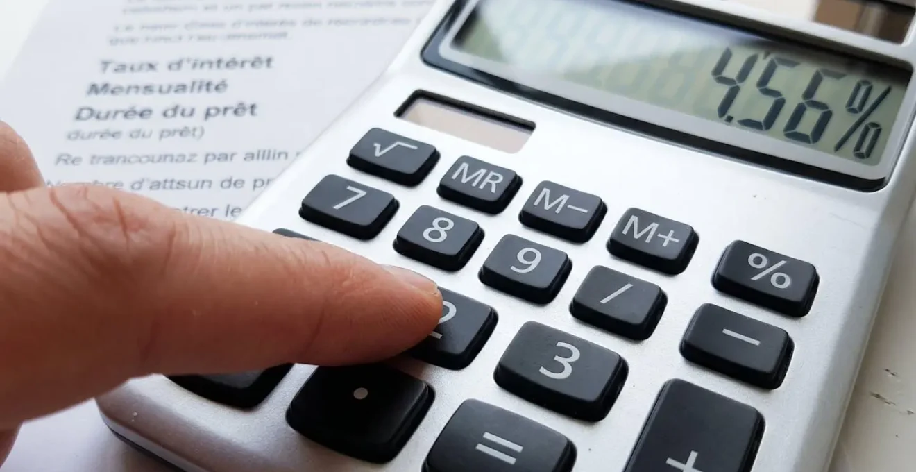 Gros plan sur une main utilisant une calculatrice moderne posée à côté d'une feuille de taux de crédit immobilier, avec un arrière-plan flouté